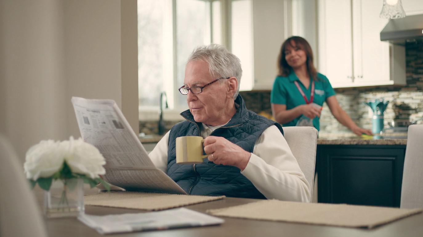 man reading newspaper with caregiver in background
